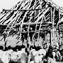 Building a simple prayer hut. Stage 2: beams and sticks have been laid over the top. Here the priest from head station addresses the catechumens.